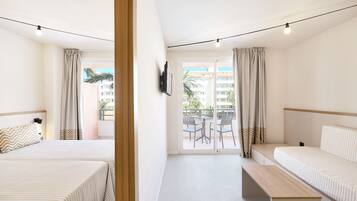 A hotel room with two beds, a bench, a TV, and a balcony with a view of buildings and palm trees.