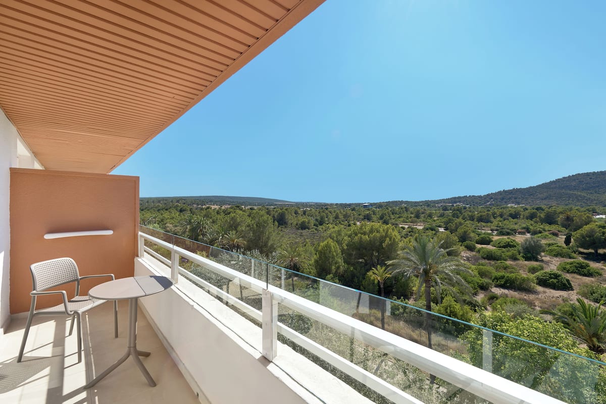 A balcony with a table and chairs, offering a view of a hilly landscape with trees and shrubs.