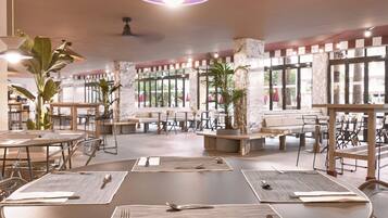 Un comedor amplio con mesas redondas, algunas con individuales y cubiertos, y ventanas grandes que dejan entrar luz natural.