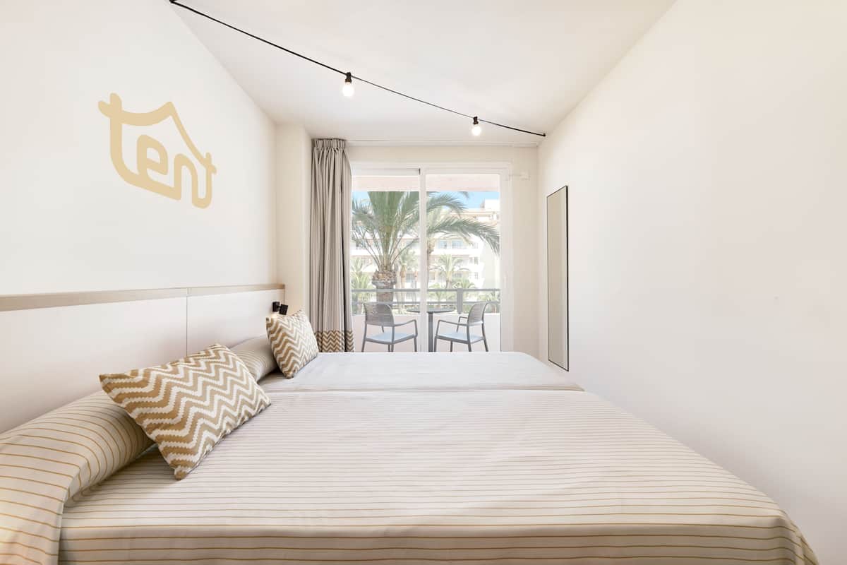 A bedroom with a bed, two chairs, a balcony with a table, and a view of palm trees and buildings.