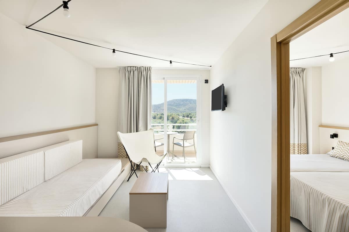 A hotel room with a bed, a sofa, a table, a TV, and a balcony with a view of the mountains.