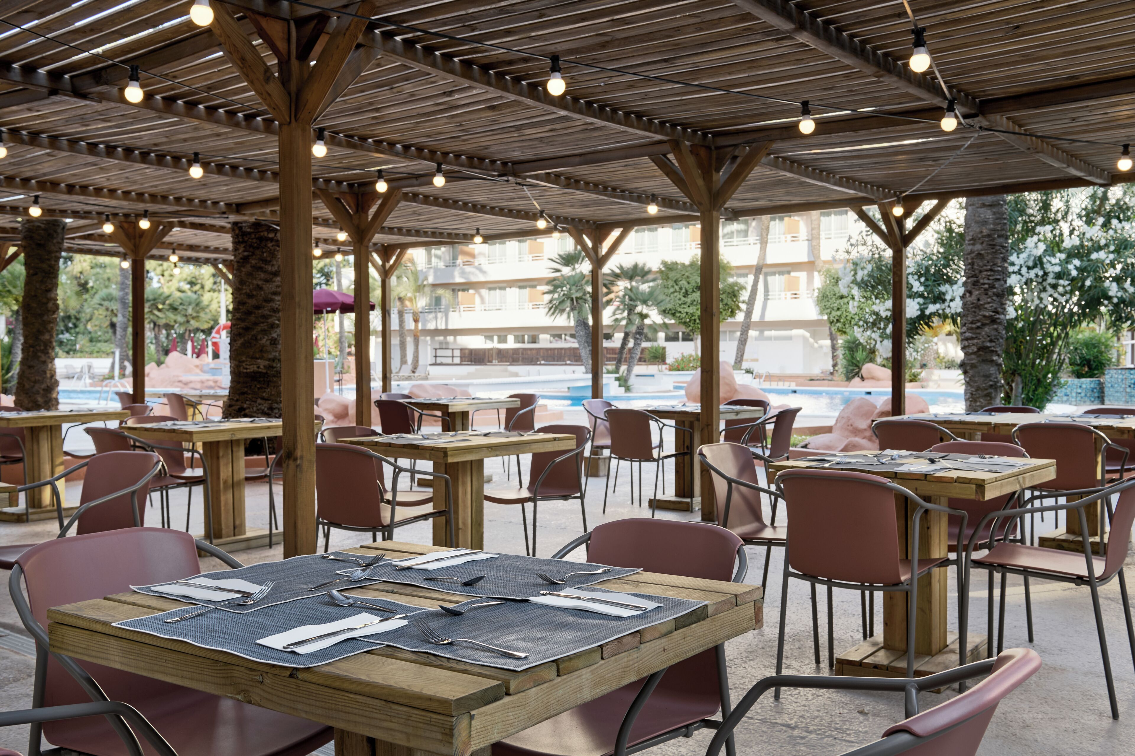 An outdoor dining area with wooden tables and metal chairs, surrounded by a covered patio with a pool view.