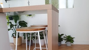 Un tabouret de bar en bois, doté d’une structure en métal blanc, placé dans une pièce au plancher en bois et agrémentée de plantes en pot.