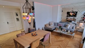 Un vaste espace de vie doté d’un parquet en bois disposé en point de Hongrie, d’une table à manger avec des chaises, d’un canapé et d’un téléviseur fixé au mur.