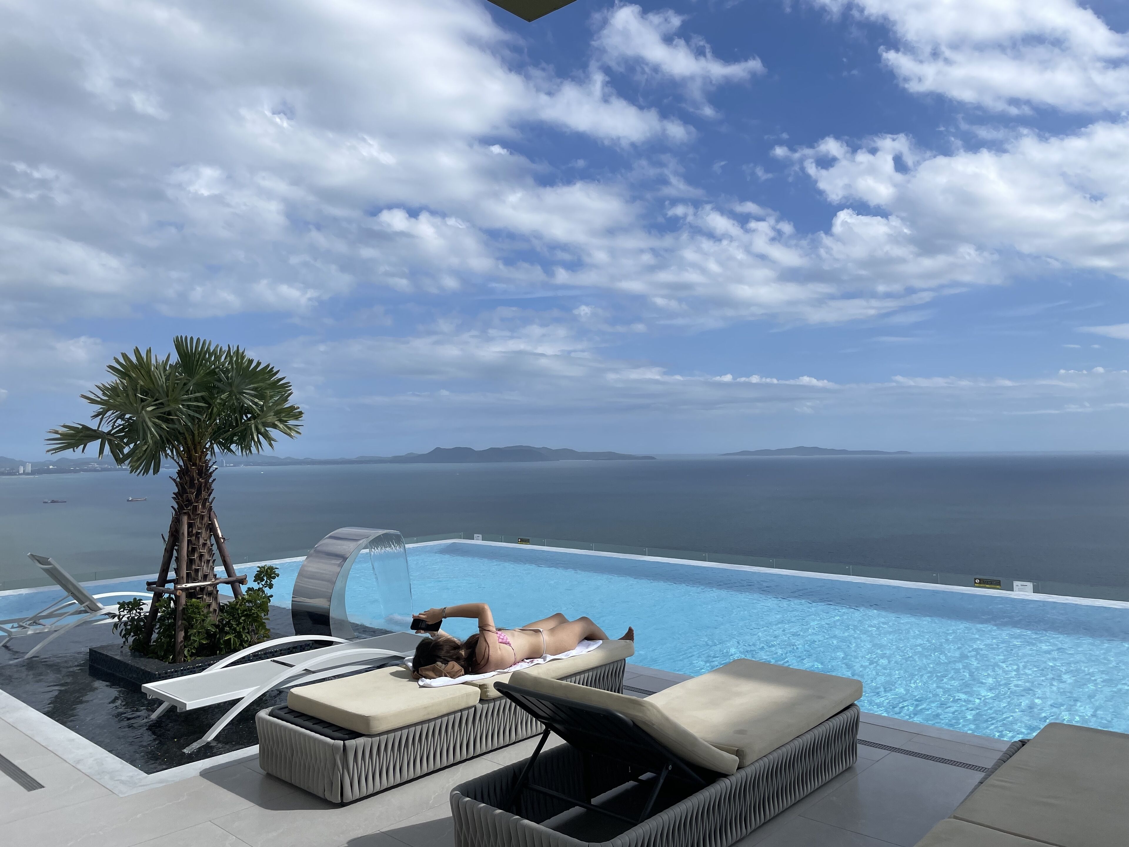 A poolside area with lounge chairs and a palm tree, overlooking a body of water and distant land.