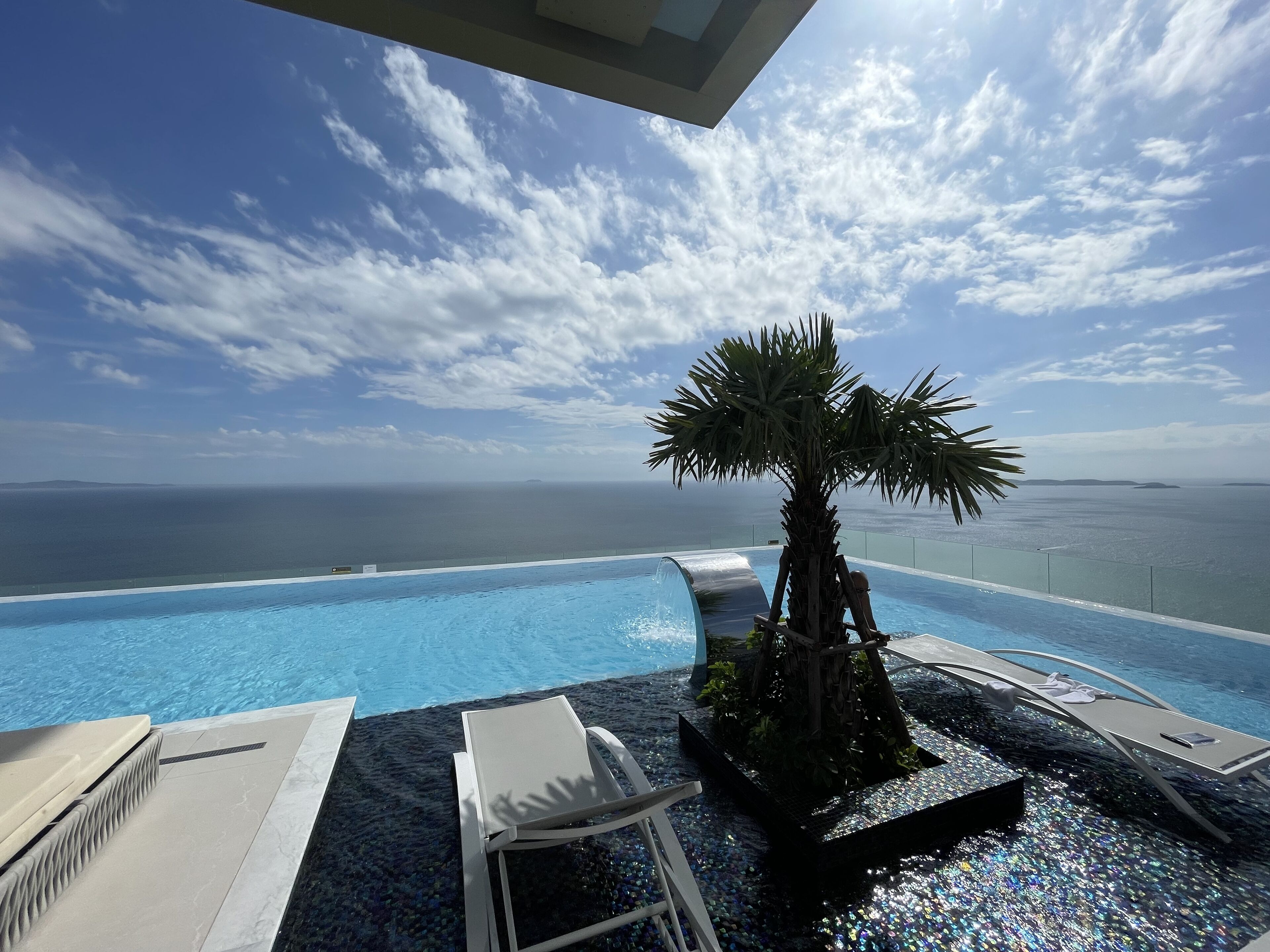 A pool area with a palm tree and lounge chairs, overlooking a body of water.