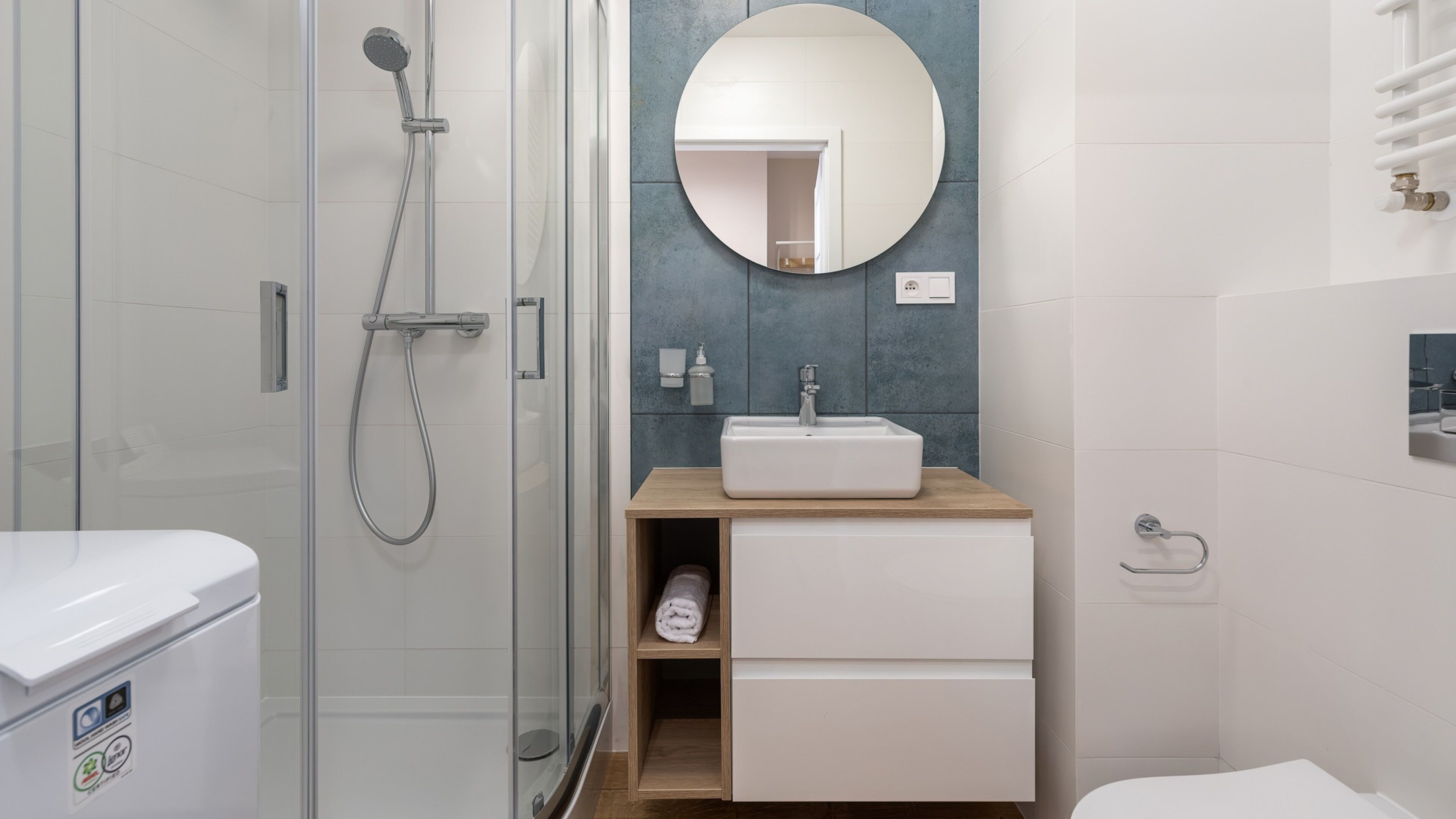 A modern bathroom with a glass shower enclosure, a wooden vanity with a sink, and a round mirror.
