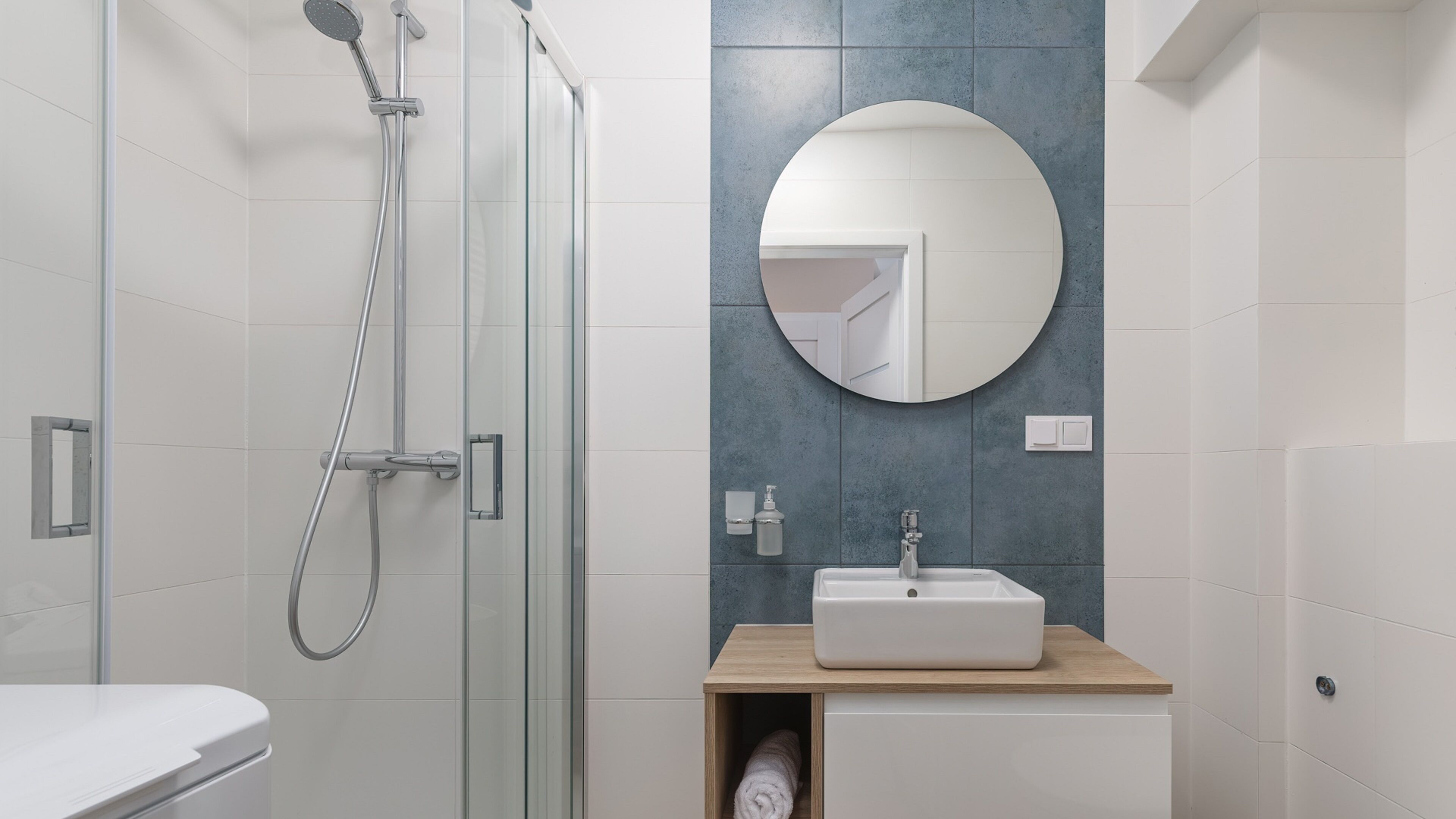 Un baño moderno con cabina de ducha de vidrio, un mueble de tocador de madera con lavamanos blanco y un espejo redondo en la pared.