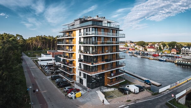A multi-story building with balconies, a waterfront view, and a parking area with cars. - Apartamenty Sun & Snow Lighthouse (Trzebiatow)