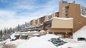 Un hôtel moderne à plusieurs étages, doté d’un toit plat et de balcons, entouré d’arbres couverts de neige et de voitures garées.