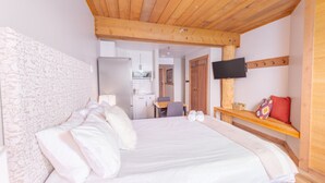 Une chambre avec un plafond en bois, un lit recouvert de linge de lit blanc, un téléviseur fixé au mur et un petit coin bureau avec une chaise.