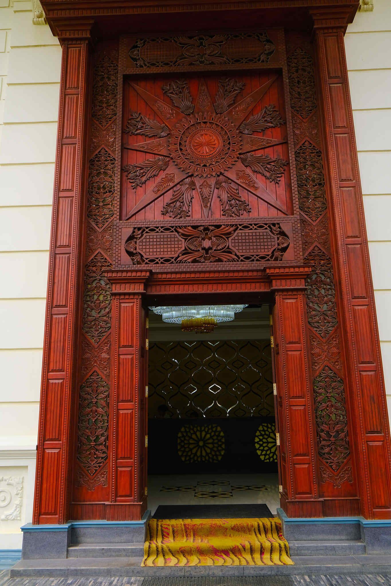A wooden entrance with intricate carvings and a yellow carpet leading up to a door with a geometric pattern.