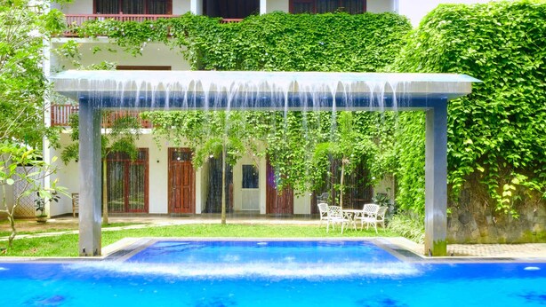 A swimming pool with a water fountain, surrounded by greenery and a building with a balcony. - Polhena Grand Resort (Matara)