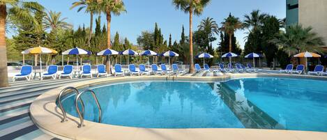 A swimming pool with blue lounge chairs and umbrellas, surrounded by palm trees and a tiled deck.