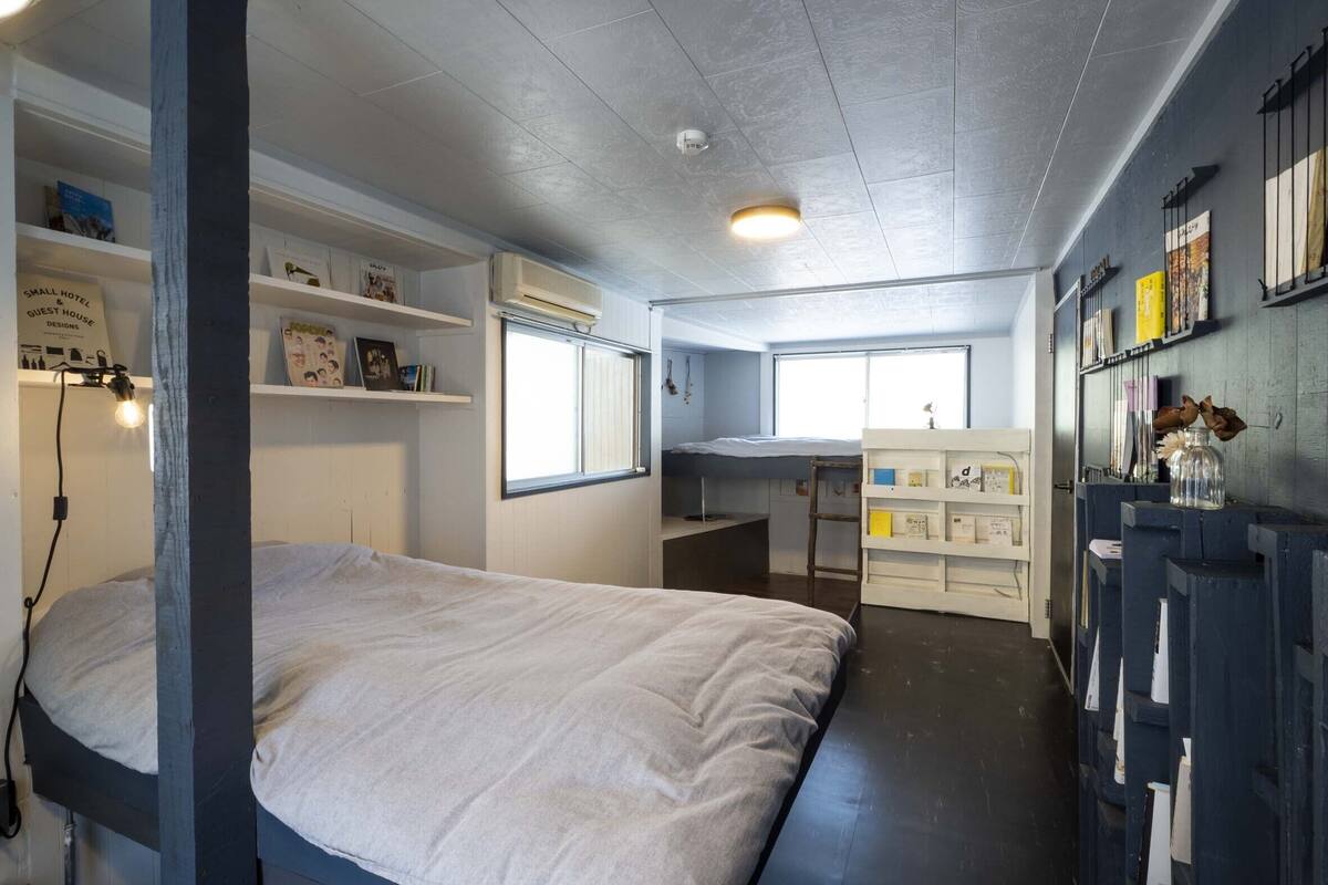 Ein kleines Hotelzimmer mit einem Bett, einem Schreibtisch, einem Regal mit Büchern, einem Fenster und einem Wandregal mit Dekoration.