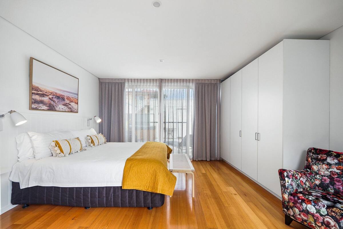 Une chambre moderne avec un grand lit, un banc, un fauteuil à motifs floraux et une vue sur l’extérieur à travers des rideaux transparents.