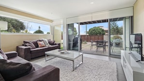 Ein modernes Wohnzimmer mit Sofa, Couchtisch und Fernseher. Es verfügt über große Schiebeglasfenster, die auf eine Terrasse mit Gartenmöbeln führen.