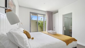 Ein modernes Hotelzimmer mit einem großen Bett, einem kleinen Tisch mit Handtüchern, einer Schiebeglastür zum Balkon und einem durch eine angrenzende Tür sichtbaren Badezimmer.