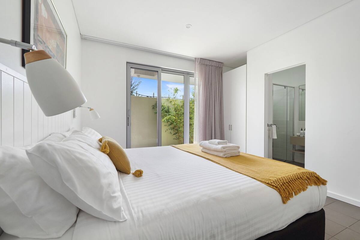 Une chambre d’hôtel moderne avec un grand lit, une petite table avec des serviettes, une porte-fenêtre coulissante donnant sur le balcon et une salle de bain visible à travers une porte adjacente.