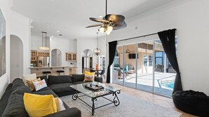 A spacious living area with a large sofa, a coffee table, and a ceiling fan. There is a sliding glass door leading to an outdoor patio with a pool.
