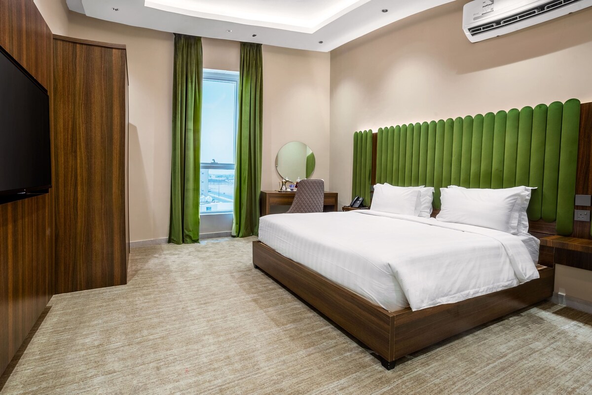 A hotel room with a large bed, a flat-screen TV, a desk with a chair, a mirror, and a view of the outside.