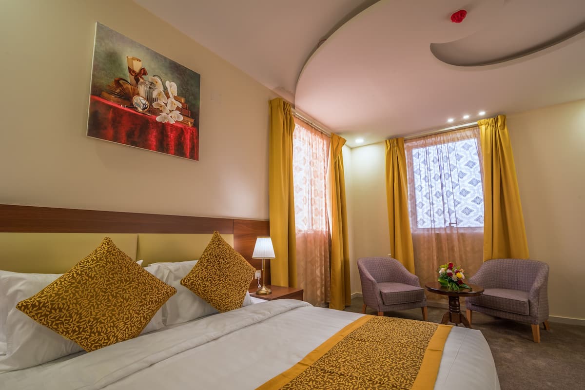 Habitación de hotel con una cama, dos sillones, una mesita con un jarrón de flores y un cuadro en la pared.