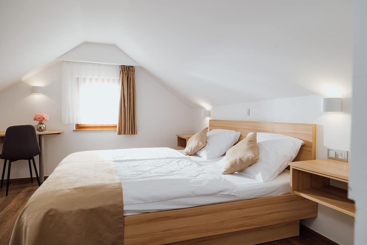 Una cama bien tendida con cabecera de madera, una mesita de noche con lámpara y un escritorio con silla.