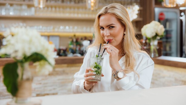 Une personne est assise à une table dans un endroit bien éclairé, à l'intérieur, et tient une boisson à la main.
