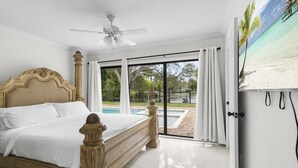 A bedroom with a large bed, a ceiling fan, a view of a swimming pool, and a beach scene on the wall.