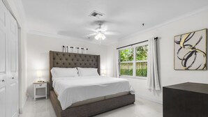 A bedroom with a large bed, a nightstand, a ceiling fan, a window with curtains, and a painting on the wall.