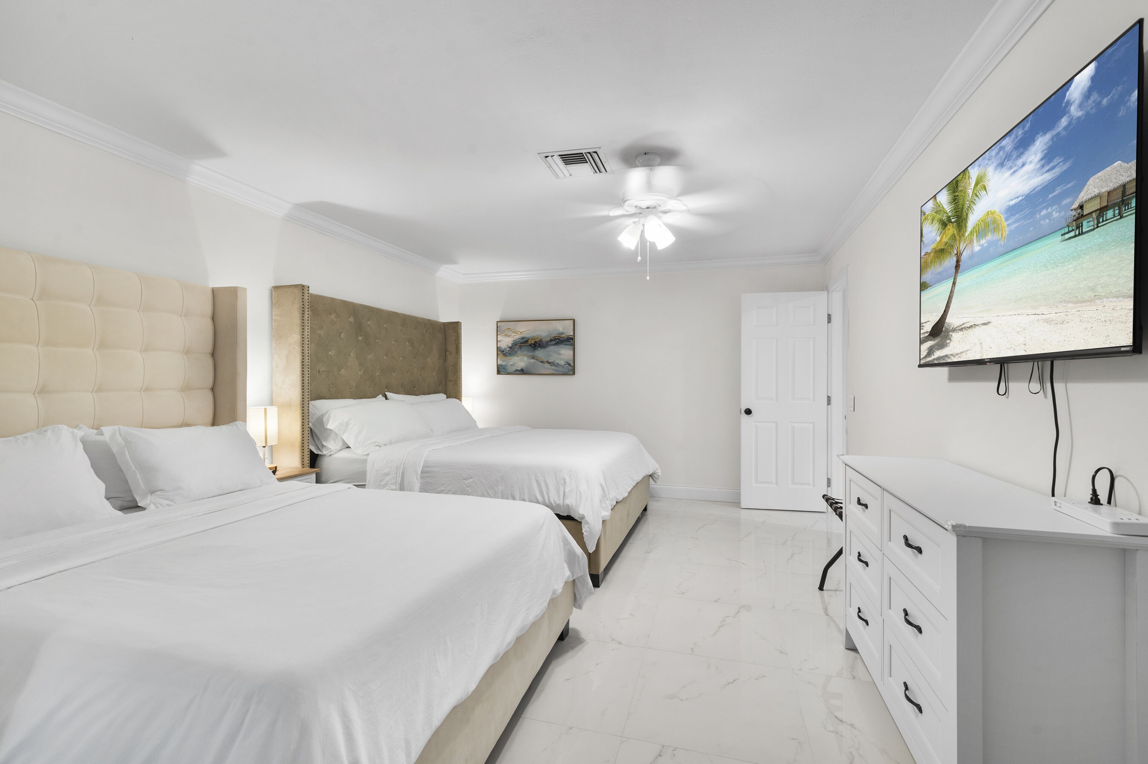 Een hotelkamer met twee bedden, een plafondventilator, een televisie en een schilderij van een strand.