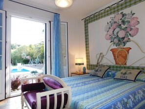 Une chambre à coucher comprenant un lit, une chaise, une table de chevet, une lampe, une fenêtre avec des rideaux, et une vue sur une piscine et la verdure environnante.