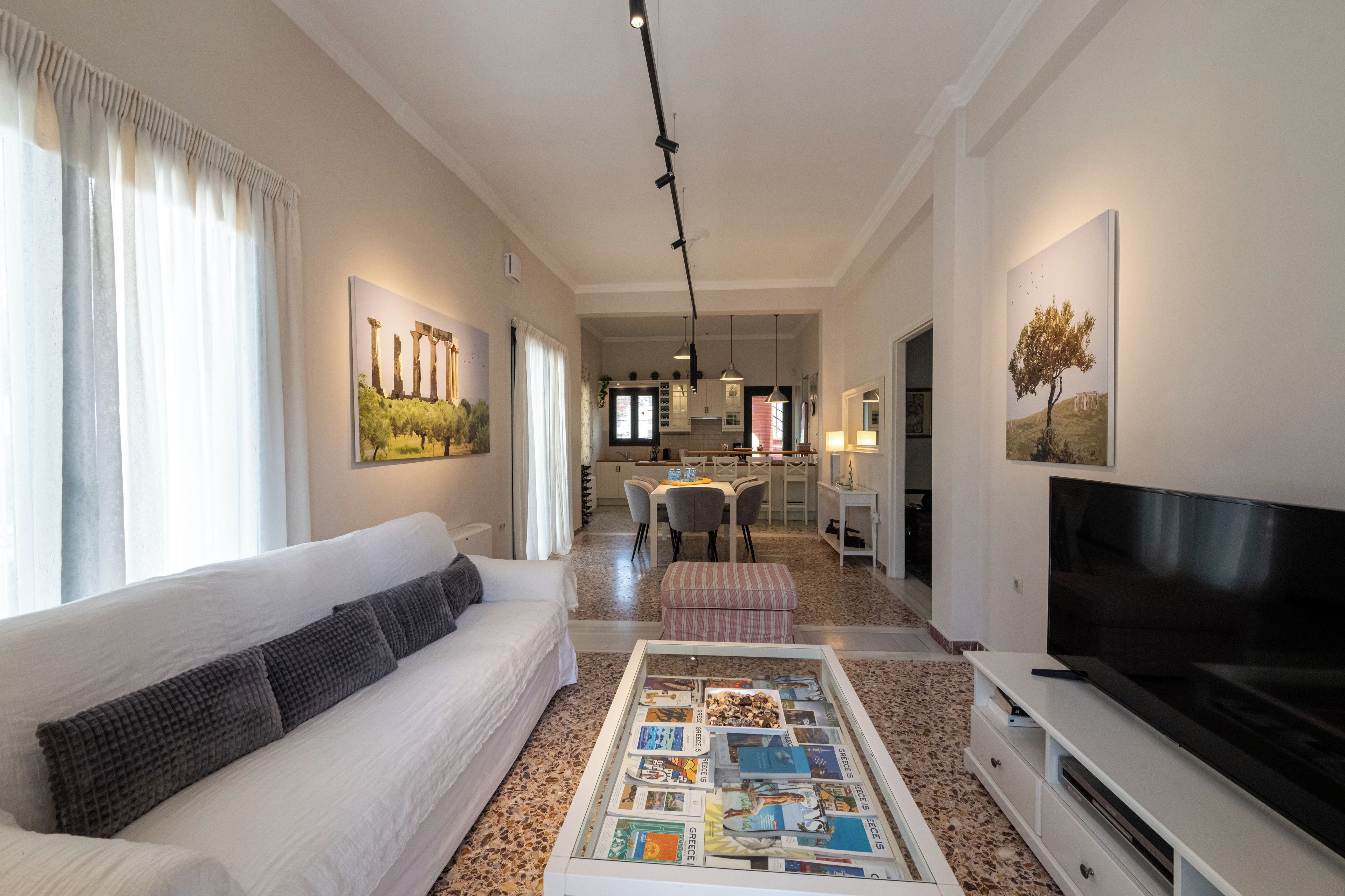 A modern living room with a white sofa, a coffee table with magazines, a flat-screen TV, and a dining area in the background.