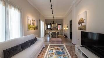 A modern living room with a white sofa, a coffee table with magazines, a flat-screen TV, and a dining area in the background.