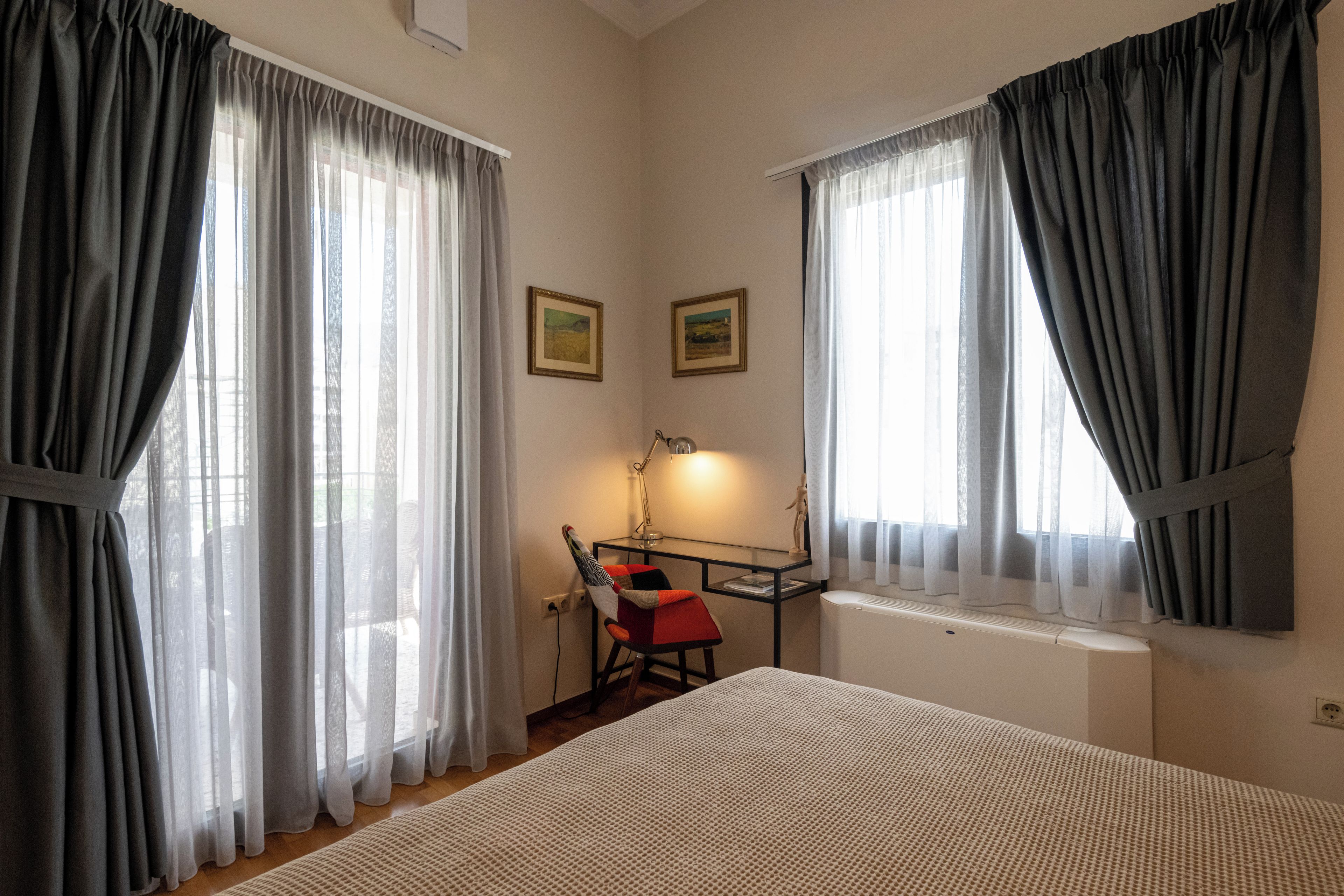 A bedroom with a bed, a desk with a chair, two framed pictures on the wall, and a window with curtains.