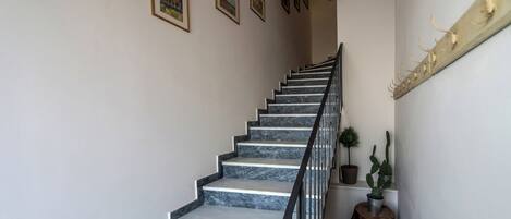 Un escalier avec des marches en marbre et une rampe en métal, menant à un palier où se trouvent un pot de fleurs et un tabouret en bois.