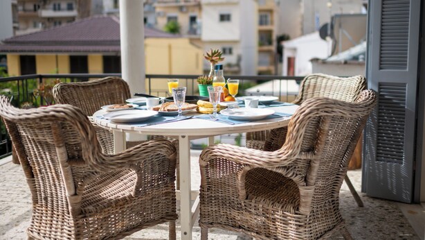 A round table with wicker chairs on a balcony overlooking a cityscape.