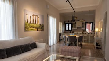 A modern living room with a sofa, a coffee table with magazines, a dining area with chairs, and a kitchen in the background.