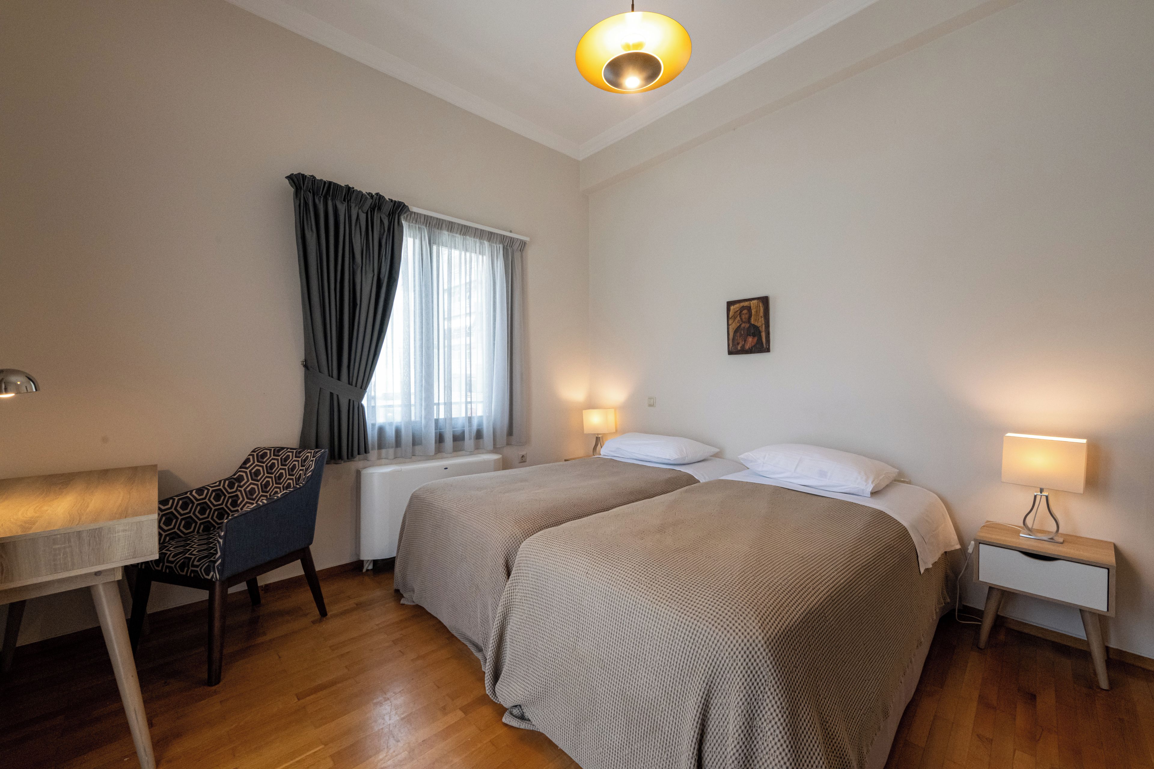 A hotel room with two beds, a desk, a chair, a bedside table, and a window with curtains.