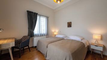 A hotel room with two beds, a desk, a chair, a bedside table, and a window with curtains.