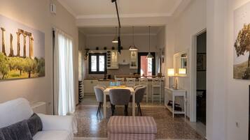 A spacious living area with a sofa, a coffee table with magazines, a dining table with chairs, and a kitchen in the background.