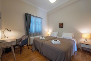Ein Hotelzimmer mit zwei Betten, einem Schreibtisch mit Lampe, einem Stuhl und einem mit Vorhängen versehenen Fenster.