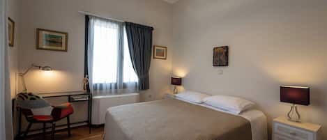 A bedroom with a large bed, a desk with a chair, a window with curtains, and two bedside lamps.
