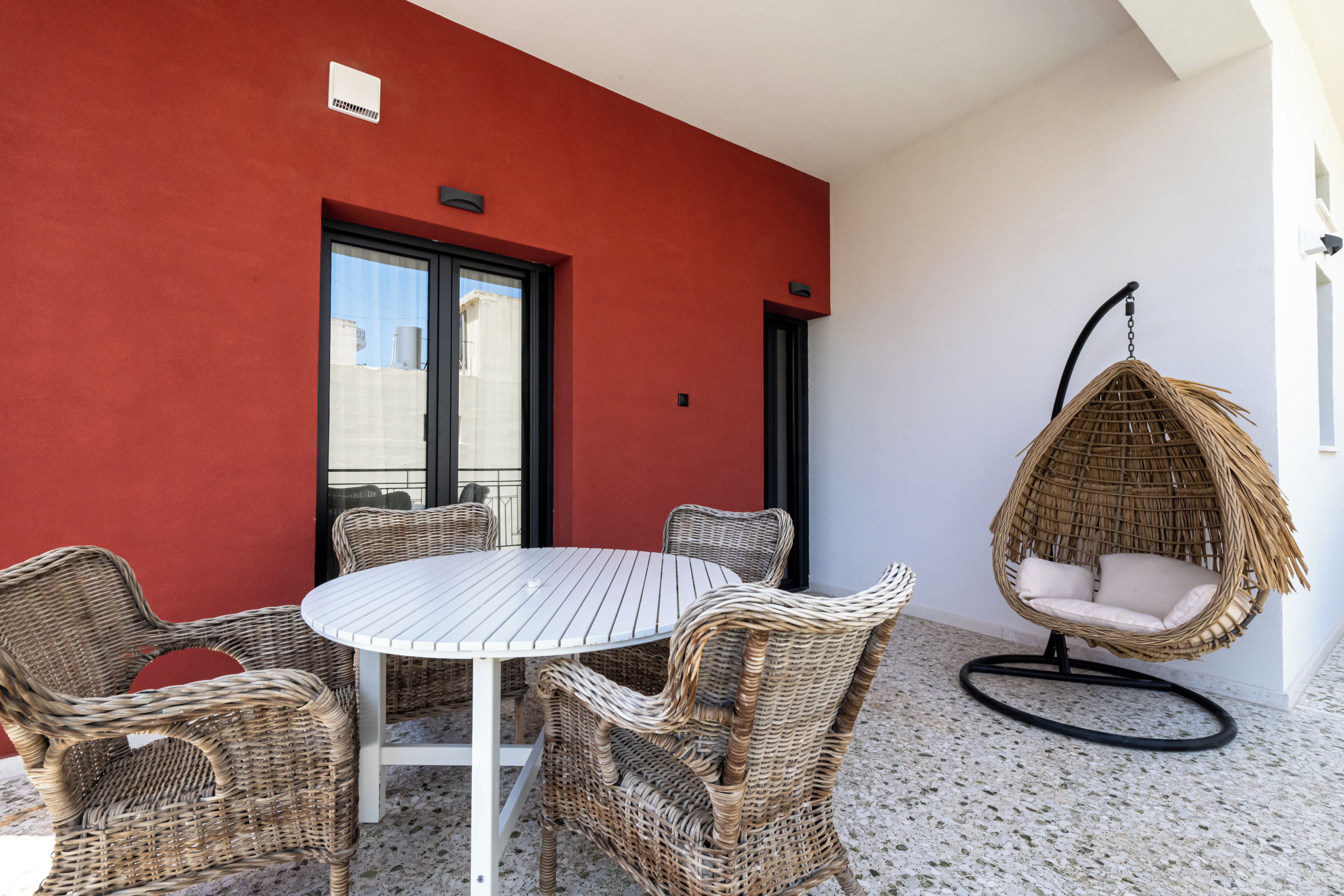 A round table with wicker chairs, a hanging chair, and a red wall with a window.