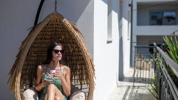 A person sitting in a wicker swing chair on a balcony with a tiled floor, overlooking a building with a white facade and a balcony railing.