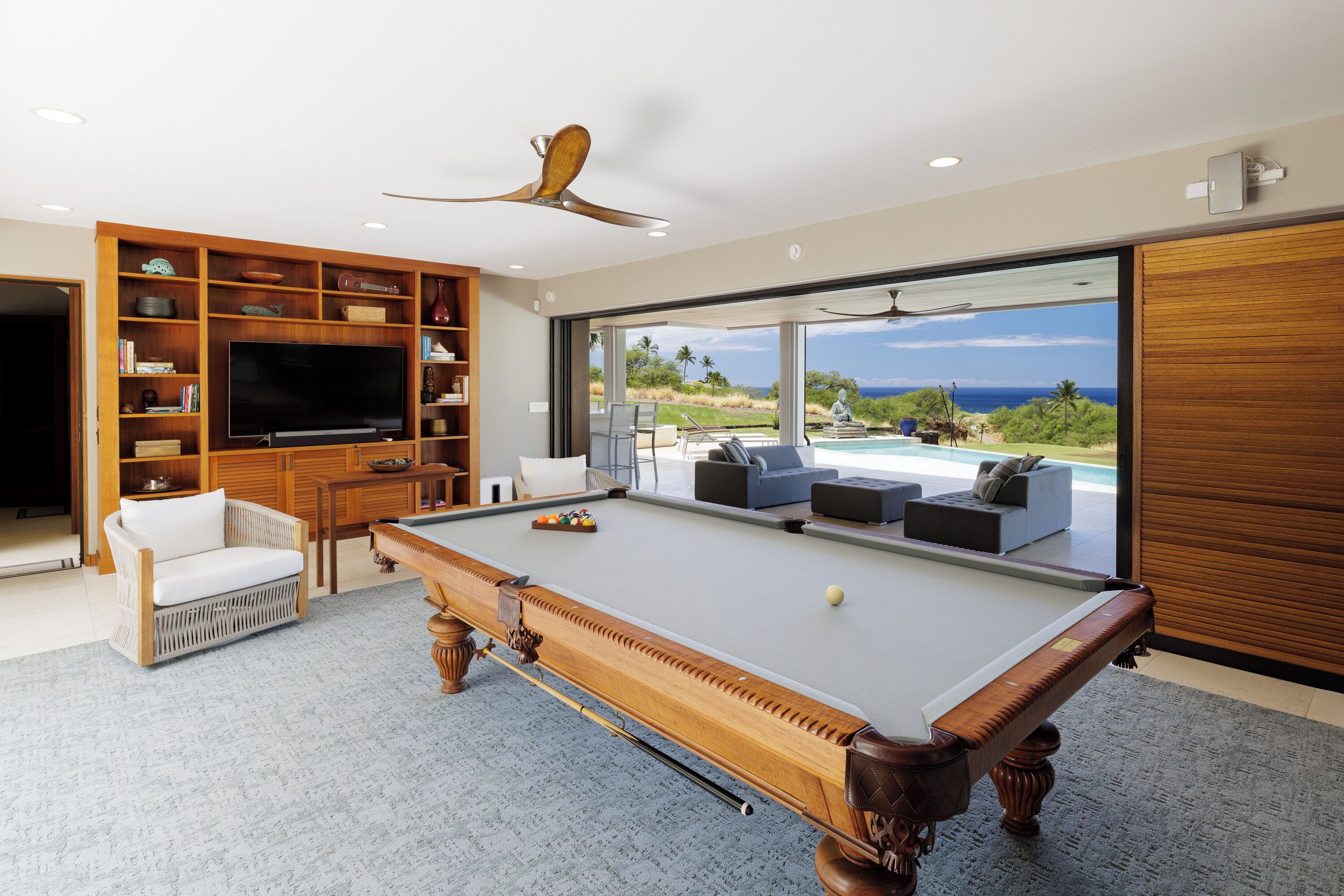 A modern living room with a pool table, comfortable seating, a television, and a large window offering a view of the outdoors.