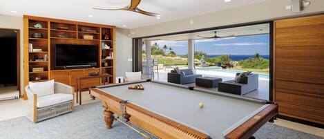 A modern living room with a pool table, comfortable seating, a television, and a large window offering a view of the outdoors.