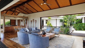 A spacious living area with a large sofa, coffee table, and chairs, wooden flooring, and a view of the outdoors. - Beautiful Mauna Kea Home, Ocean and Sunset Views (Kamuela)