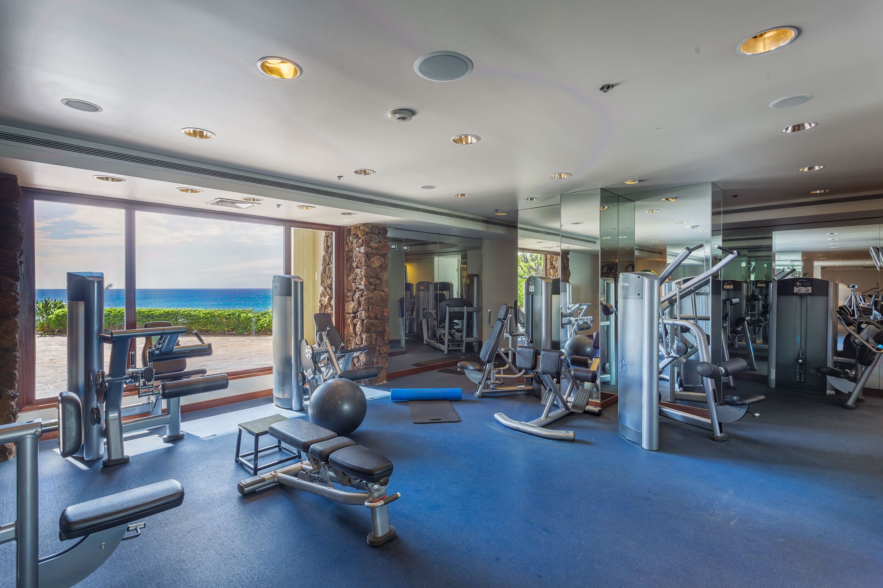 Ein Fitnessraum mit diversen Trainingsgeräten, großen Fenstern mit Meerblick und einer Steinsäule.