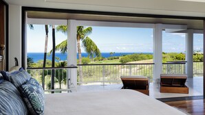 A bedroom with a large bed, a balcony with a view of the ocean and palm trees, and a wooden bench. - Beautiful Mauna Kea Home, Ocean and Sunset Views (Kamuela)